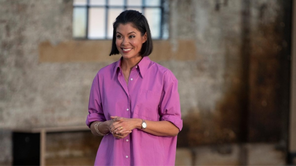 Journalist Alex Wagner in a relaxed professional pose, wearing a purple shirt and gold accessories in an industrial-style setting.