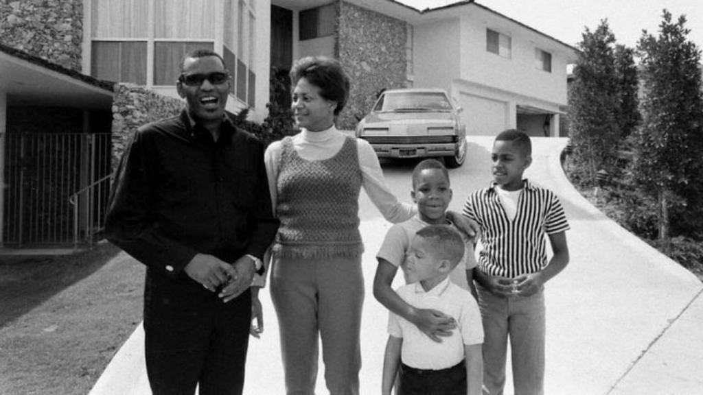 A nostalgic black and white vintage photo of Ray Charles and Della Bea Robinson standing with their three sons in the driveway of their mid-century home.