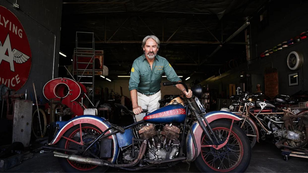 Mike Wolfe standing inside Antique Archaeology surrounded by vintage signs and motorcycles.