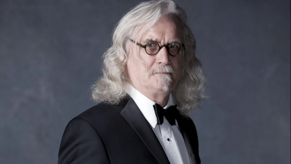 Sir Billy Connolly, Jamie Connolly’s father, posing in a colorful jacket and signature glasses at a media event.